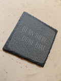 A square dark gray slate coaster with a textured surface and the phrase 'BEAN THERE DONE THAT' debossed on top, resting on a light wood surface.