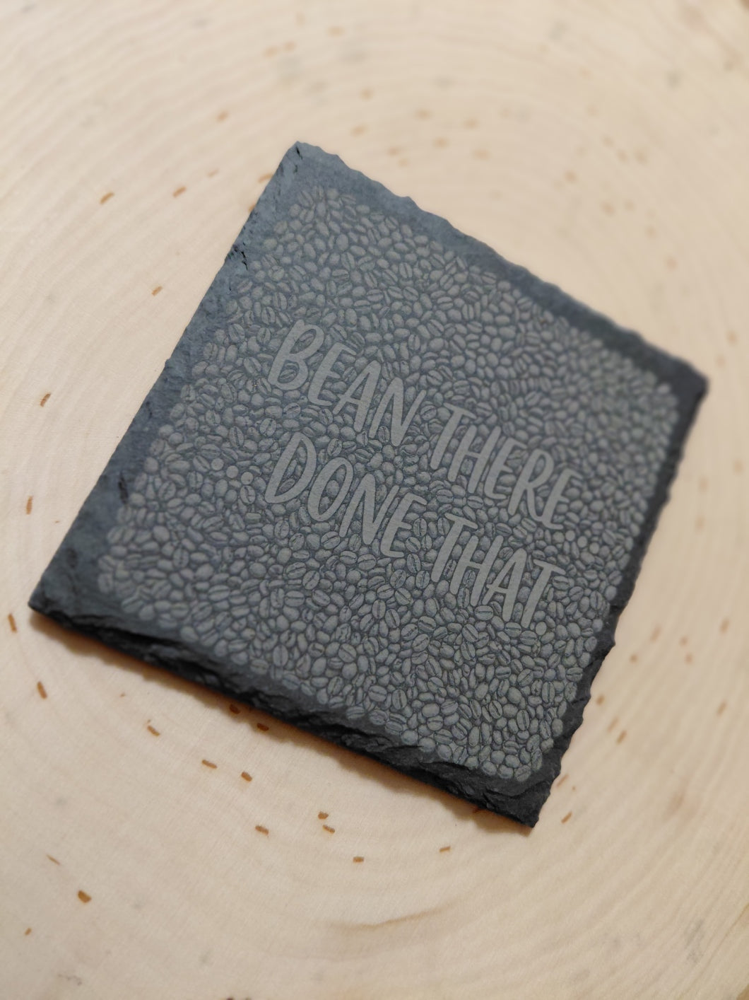 A square dark gray slate coaster with a textured surface and the phrase 'BEAN THERE DONE THAT' debossed on top, resting on a light wood surface.